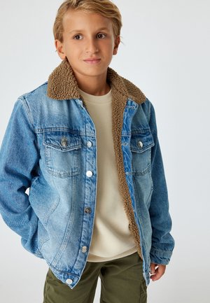 Jeansjakke med en brun shearlingkrave, to brystlommer og sølvfarvede detaljer. Bæres over en lys beige sweater, parret med grønne bukser.