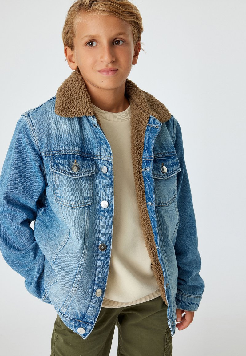 Jeansjacka med en brun fårskinnskrage, två bröstfickor och silverdetaljer. Bärs över en ljusbeige tröja, tillsammans med gröna byxor.