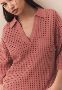 Rožinis megztas megztinis su V formos iškirpte ir trumpomis rankovėmis, pasižymintis tekstūruotu atviru audimu ir apykakle.