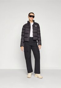 Svart puffjacka med hög krage och dragkedja, kombinerad med svarta cargobyxor. Modellen bär mörka solglasögon och beige sneakers.