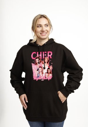 Kobieta w czarnej bluzie z kapturem z różowym napisem „CHER” i obrazkami czterech kobiet, pod spodem tekst „Ugh! As if!”, uśmiechnięta i patrząca w bok.