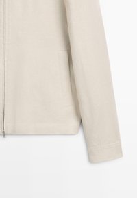 Beiger Jackenärmel und unterer Torso mit Seitentaschendetail und vorderem Reißverschlussverschluss vor einfarbigem Hintergrund.