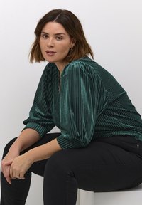 Grøn plisseret bluse med lange puffærmer og v-hals, lavet af skinnende materiale. Båret med sorte bukser, siddende mod en ensfarvet baggrund.