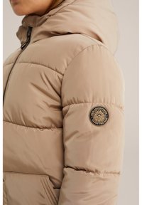 Doudoune beige avec capuche, présentant des sections matelassées horizontales, une fermeture éclair à l'avant et un patch rond noir sur la manche gauche.