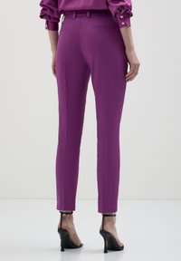 Persona in pantaloni slim-fit viola e camicia abbinata, abbinati a sandali neri con tacco alto, in piedi davanti a uno sfondo neutro.