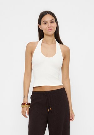 Femme aux longs cheveux foncés portant un haut blanc à col halter et un pantalon ample foncé, debout avec une expression détendue sur un fond uni.