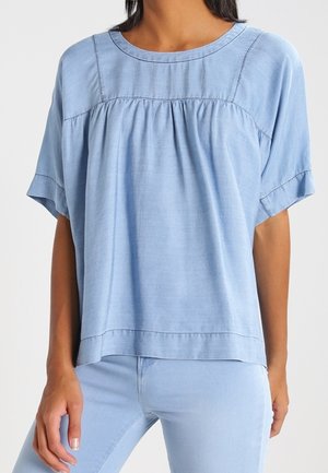 Kobieta w luźnej, krótkim rękawem, jasnoniebieskiej bluzce z marszczonym karczkiem, w połączeniu z dopasowanymi jasnoniebieskimi spodniami.