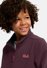 Veste en polaire violette avec un col haut, arborant un logo rose sur la poitrine et une fermeture éclair à l'avant. Texture douce, design bien ajusté.