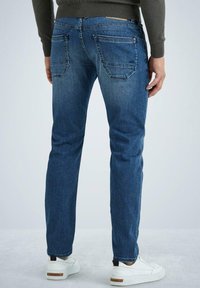 Jeans de mezclilla azul oscuro con corte recto, que cuentan con dos bolsillos traseros y detalles de desgaste sutil. Llevados con zapatillas blancas.