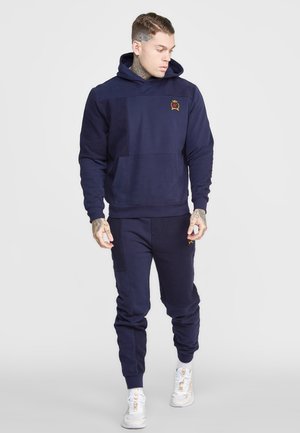 Mörkblå hoodie och jogging-set i mjukt tyg, med framficka, applikationsdesign och broderad logga på bröstet.