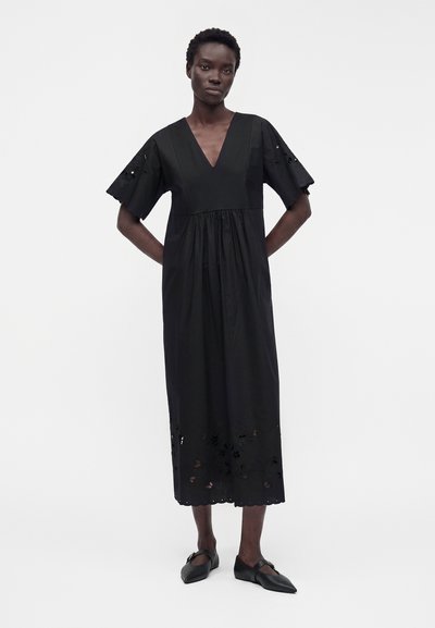 Mannequin portant une robe midi noire avec col en V, manches courtes larges, détails découpés floraux sur les manches et l'ourlet, associée à des chaussures plates noires.