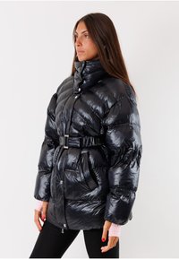 Giacca nera in piuma con colletto alto, vita cinturata e finitura lucida. Presenta grandi sezioni trapuntate e tasche. Maniche lunghe con polsini rosa chiaro.