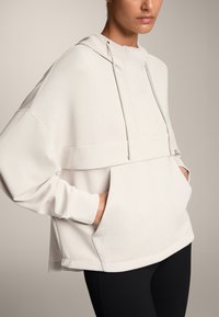 Un hanorac supradimensionat de culoare bej deschis, cu un buzunar frontal, șnururi și o textură netedă. Designul include un stil scurtat și mâneci largi.