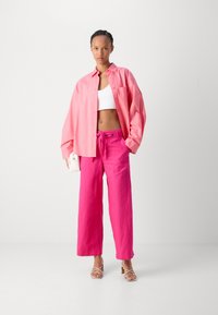 Rosa oversized skjorte, hvit crop top og lys rosa vide bukser; modell som har på seg strappy hæler, holder en liten hvit veske.