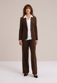 Bruin op maat gemaakt pak met een getailleerd colbert en broek met wijd uitlopende pijpen, gecombineerd met een wit overhemd met kraag, aangevuld met zwarte pumps.