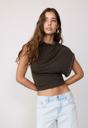 Ung kvinde med langt, bølget hår iført en olivengrøn asymmetrisk top med metaløjer og lyseblå jeans med højtaljet pasform.