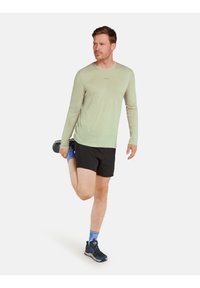 Hellgrünes langärmliges Sportshirt, schwarze Shorts, blaue Laufschuhe, blau gemusterte Socken. Mann dehnt sein Bein im Stehen.