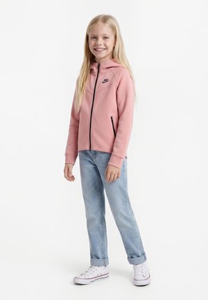 Noor tüdruk, kes naeratab, kannab roosat Nike lukuga kapuutsi, helerohelisi üleskeeratud pükse ja valgeid tossusid, taustal valge sein.