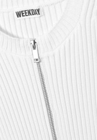 Haut blanc côtelé à fermeture éclair avec un col rond. Dispose d'une fermeture éclair argentée et d'une étiquette portant l'inscription "WEEKDAY" près du col.