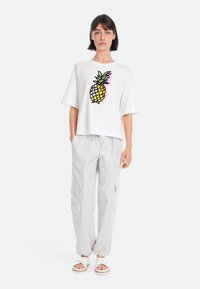 White oversized t-shirt featuring a colorful pineapple graphic. Paired with light grey cargo pants and white sandals. Simple design and casual fit.