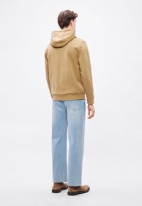 Béžová mikina s kapucňou na sťahovanie, kombinovaná s svetlomodrými džínsami s rovnými nohavicami. Hladká látka s rebrovanými manžetami a lemom, nosená s hnedými topánkami.