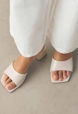 Feet wearing white open-toe block heel sandals paired with white wide-leg pants on a neutral floor.