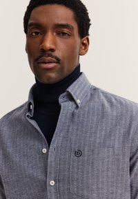 Uomo con capelli ricci corti e baffi indossa una camicia grigia a spina di pesce sopra un dolcevita nero, su uno sfondo chiaro e uniforme.