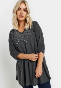 Grijze V-hals top met driekwart mouwen, voorzien van kralendetails en gathers op de mouwen, gemaakt van zachte, lichte stof.