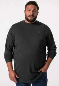 Langärmliges, dunkelgraues T-Shirt aus weichem Material mit abgerundetem Saum und subtiler Logo auf der linken Brust. Kombiniert mit blauen Jeans.