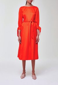 Femme portant une robe midi rouge vif avec des manches longues attachées aux poignets et une ceinture à la taille, assortie à des talons pointus couleur nude.