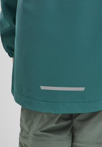 Giacca teal con polsini elasticizzati, orlo dritto e una striscia riflettente grigia nella parte inferiore. Il tessuto appare liscio e leggero.
