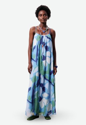 Femme aux cheveux courts et bouclés portant une robe longue sans manches à motifs bleus, verts et blancs, avec des colliers perlés superposés et des chaussures vertes.