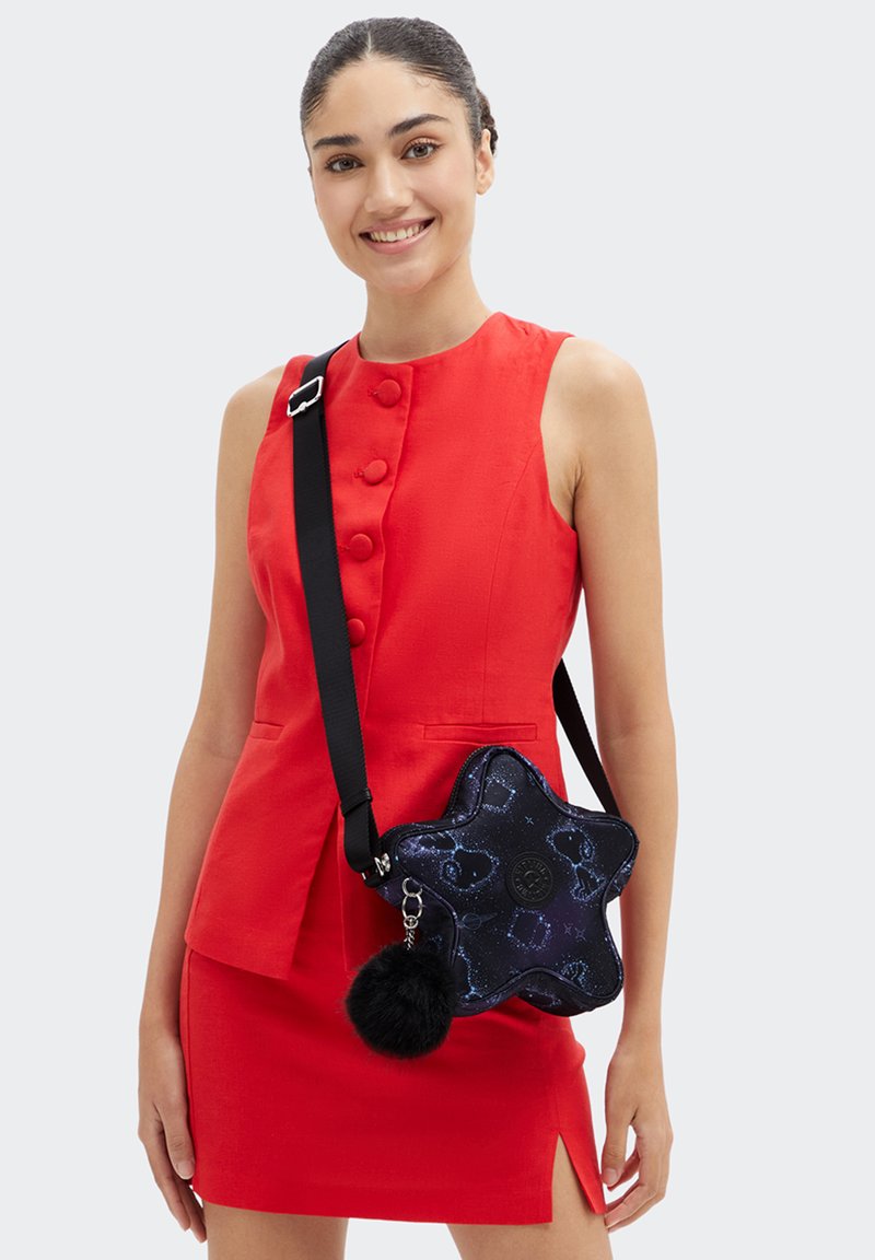 Ung kvinde, der smiler, iført en ærmeløs rød kjole og en sort stjernesvetteret crossbody taske med en fluffy pom-pom charme.