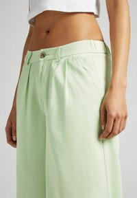 Light green trousers with a high waist, pleated design, and a button closure. Features subtle branding in thread on the side.