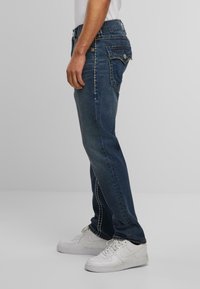Vaqueros de denim en un lavado azul oscuro con costuras en contraste, un diseño de pierna recta y bolsillos traseros adornados. Zapatillas blancas puestas.