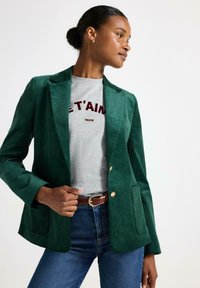 Zelený kordový blazer se dvěma náprsními kapsami a zlatými knoflíky. Stylizováno přes šedé tričko s červeným textem, v kombinaci s modrými džíny a hnědým opaskem.