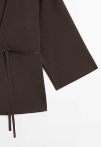 Donkerbruin langmouwig omslagvest met geribbelde textuur en een smalle strikceintuur in de taille, afgebeeld op een witte achtergrond.