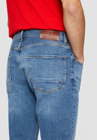 Jeans en denim bleu clair avec une patch en cuir au dos, des poches cousues et une petite étiquette logo. Coupe droite avec des coutures visibles.