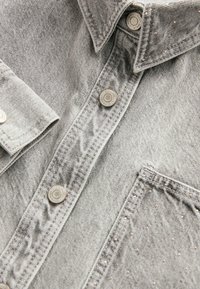 Chemise en jean gris clair avec boutons-pression métalliques et petits clous décoratifs sur le col et la poche.