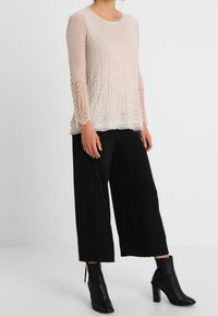 Ljusbeige långärmad topp med texturerad stickning och spetskant, kombinerad med svarta vida byxor och ankelstövlar. Platt yta, neutral bakgrund.