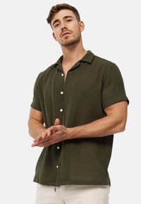 Camicia a maniche corte di colore verde oliva, con bottoni in tessuto testurizzato, dotata di colletto e bottoni bianchi, indossata con pantaloni chiari.
