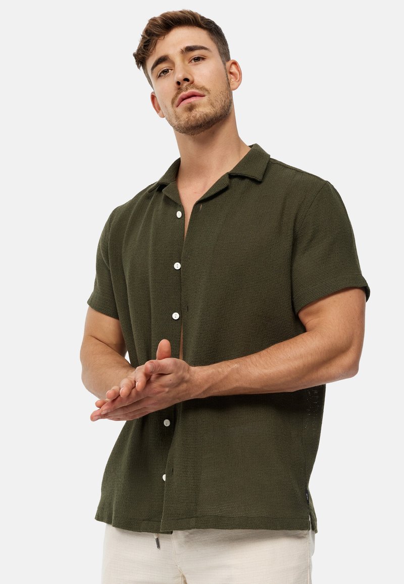 Camicia a maniche corte di colore verde oliva, con bottoni in tessuto testurizzato, dotata di colletto e bottoni bianchi, indossata con pantaloni chiari.