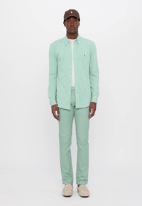 Lichtgroen overhemd met knopen, lichtgroene broek en beige schoenen. Bruine pet met logo aan de voorkant. Glad stoftextuur, nauwsluitende pasvorm.