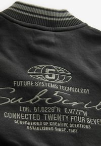 Svart sweatshirt-design med ribbad krage i grå ränder och broderad text som detalj på teknik och platskoordinater.