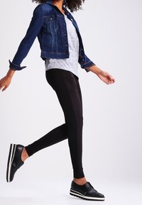 Veste en jean avec délavage bleu, t-shirt gris, leggings noirs et chaussures noires avec bandes blanches. Mannequin dans une pose dynamique.