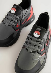 Grå og sorte træningssko med røde accenter, med et Poké Ball-logo på tungen, tekstureret stof og snørebåndsdesign.