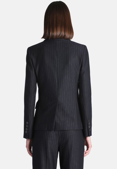 Personne aux cheveux bruns et raides portant une veste de costume sombre à fines rayures et un pantalon assorti, vue de dos sur un fond uni.