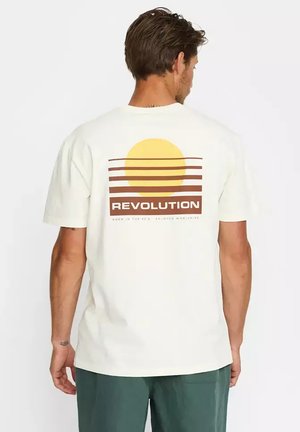 Hombre con una camiseta blanca que muestra un sol amarillo y rayas marrones con la palabra "REVOLUTION" en la espalda, combinada con pantalones verdes.