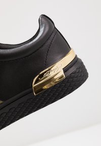 Zapatilla de cuero negra con suela texturizada negra y acento metálico dorado en el talón grabado con el nombre de la marca.