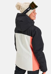 Giacca da sci con cappuccio nero, design a blocchi di colore arancione e bianco, materiale impermeabile e berretto lavorato a maglia con pompon.
